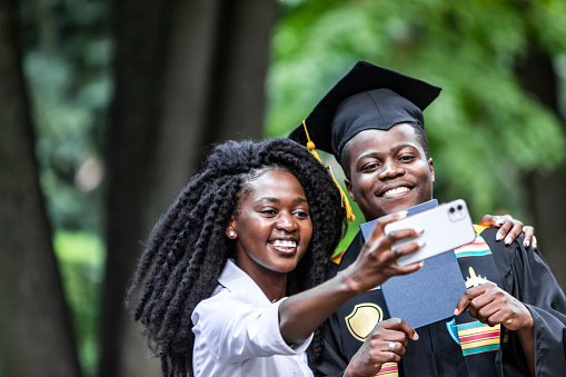 Young black woman with black male university graduate wearing black gown and cap making selfie using mobile