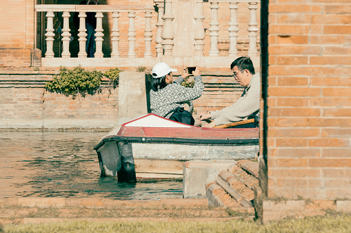 Seville, Spain; January 25th 2024: Asian tourists paddling a small boat in the canal of the Plaza de Espaa. A woman takes a selfie while her husband rows.