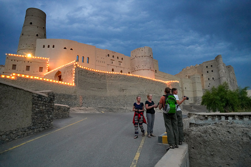 Evening over Tourists looking and photographing ruins of historic village with Bahla Fort with lights in background, Oman.