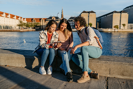 Three young friends using smart phone while sitting on the dock in Copenhagen in Denmark.