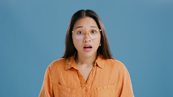 Portrait, wow and surprise of Asian woman with news, announcement or gossip. Face, shock and amazed person in glasses with secret, wtf and omg emoji isolated on a blue studio background mockup space