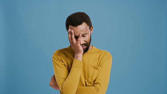 Black man, tired and disappointment in studio with worry, face palm and memory of mistake. Male person, upset and ashamed with regret for decision, isolated and blue background with headache