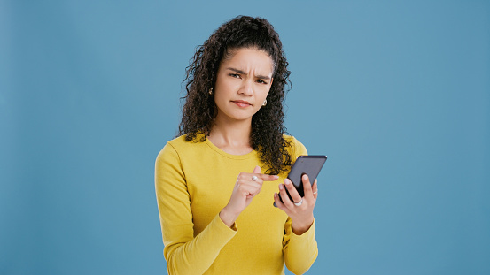Confused, woman and portrait with phone for news, announcement and doubt information in studio. Doubt, why or girl frustrated by spam notification on smartphone with error glitch on blue background