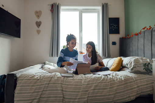 Two girls sitting together on a bed in a bedroom doing homework at their home in the North East of England. They are using a digital tablet to help them with the work. The older girl is tutoring the younger one, mentoring her in her studies.
