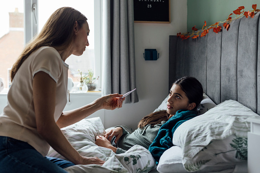 Young girl laying in her bed feeling unwell at her home in the North East of England. Her mother is checking up on her, reading from a temperature thermometer.