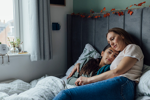 Young girl laying in her bed feeling unwell at her home in the North East of England. Her mother is checking up on her, sitting with her around her in an embrace.