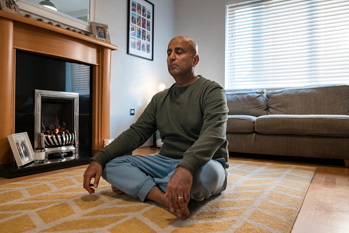 Man meditating, practising mindfulness in his living room at home in the North East of England. He is sitting crossed legged on the floor with his eyes closed.