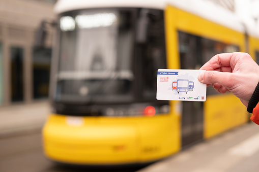 male hand hold electronic Deutschland-Ticket, yellow tram Flexity Berlin driving down city street in Berlin, monthly travel pass in germany for public transport, Berlin, Germany - February 19, 2024