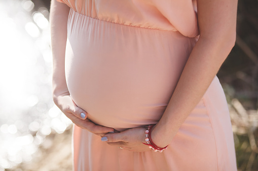 Close up photo of young pregnant woman in elegant pink dress touching her belly with hands