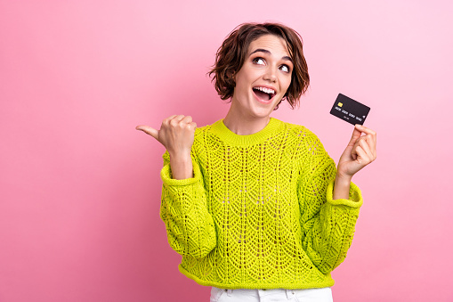 Photo of cute young girl shopaholic wear yellow knitted sweater directing finger mockup with e card isolated on pink color background.