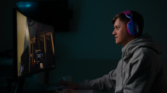 Tracking shot of happy gamer male in headphones playing first-person shooter online video game on powerful personal computer sitting at desk in dark room.