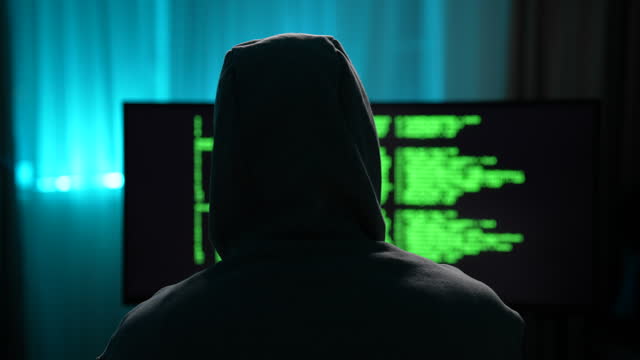 Rear view of unrecognizable male hacker wearing sweatshirt with hood typing on keyboard looking to monitor with green text code on screen. Computer hacker writing code