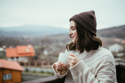 Young woman in white knitted sweater holding white cup, drinking hot tea, standing on a balcony of a cozy wooden house with mountains view.