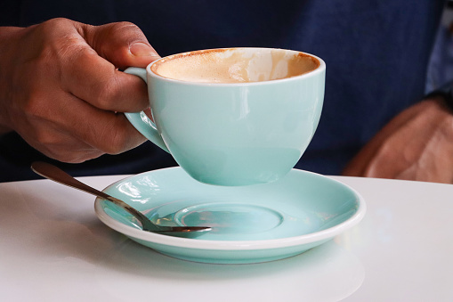 Stock photo showing close-up view of an unrecognisable man picking up a cup of cappuccino coffee from a pale blue saucer with a metal teaspoon. The freshly brewed coffee is being served on a table at a cafe, a hot energising beverage ready to drink.