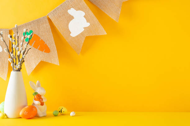 istock Celebrate Easter mood. A side view of a table adorned with figurine bunny with carrot, multicolored eggs, pussy willow in a vase, and a farmhouse garland on an orange backdrop with space for wording 2060685063