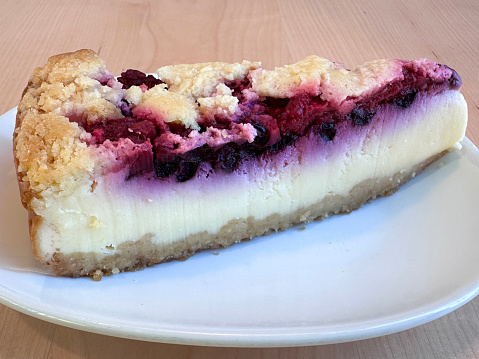 Stock photo showing close-up view of blackcurrant cheesecake with buttery biscuit base on ceramic plate, against a restaurant table background.