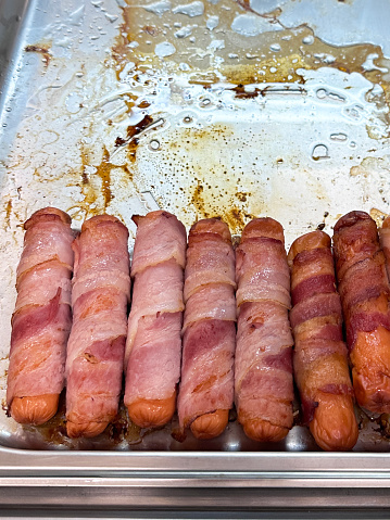 Stock photo showing close-up, elevated view of a metal serving tray containing sausages wrapped in bacon (pigs in blankets) for a breakfast self-service buffet at a hotel, to be eaten as part of a greasy and filling meal.
