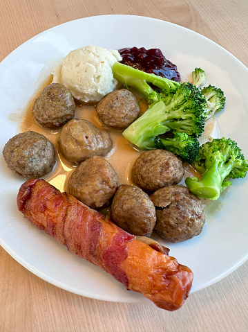 Stock photo showing close-up, elevated view of Swedish meatballs served with gravy, sausage wrapped in bacon, broccoli, scoop of mashed potato and cranberry sauce on a white plate.