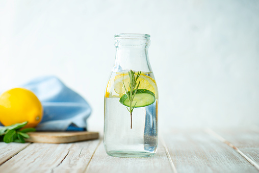 Bottle of infused water on white wood with a slice of lemon , cucumber and rosemary leaf in it.