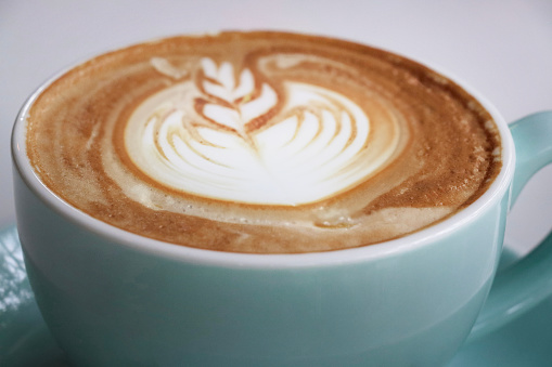 Stock photo showing close-up view of a cappuccino coffee served for breakfast on a cafe table. The cappuccino has a typically frothy milk foam surface with a flower and leaves pattern latte art design.