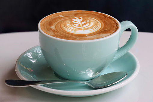 Stock photo showing close-up view of a cappuccino coffee served for breakfast on a cafe table. The cappuccino has a typically frothy milk foam surface with a flower and leaves pattern latte art design.