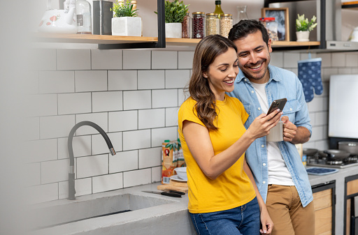 Latin American couple at home looking at social media on their cell phone while having a cup of coffee in the kitchen â lifestyle concepts