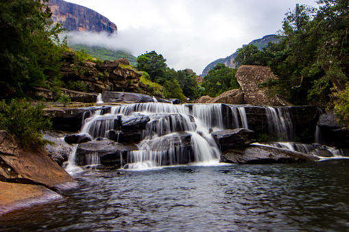 The Mahai cascades in the early morning, with the Drakensberg Mountains shrouded in mist in the background. The Cascades is a famous small waterfall and swimming spot in the Mahai river in the Royal Natal National Park in the Drakensberg Mountains of South Africa.