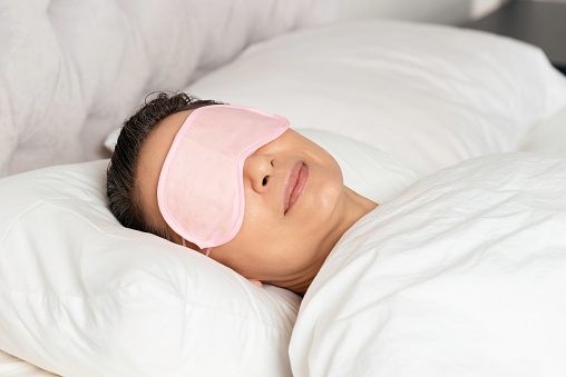 Sleeping caucasian female with pink eye mask and a warm smile in white bed.