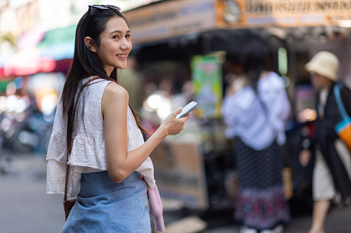 Attractive young Asian tourist is walking on the street in a retail market in Thailand. She is using a smartphone to send a message and is calling someone on the mobile phone.