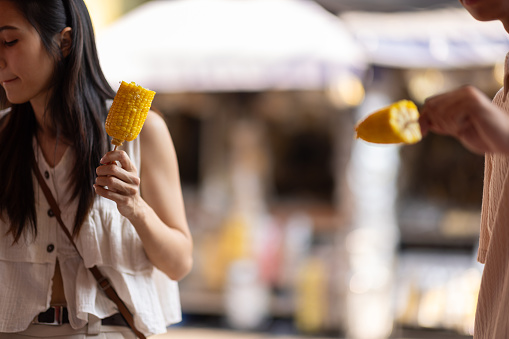 Young Asian woman holding boiled sweet corn eating while walking on street food market in Thailand.