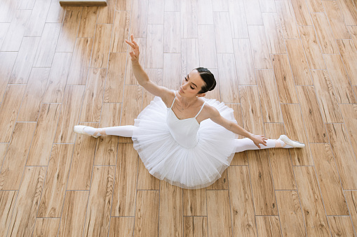 Young woman ballerina in white pack posing on background of wooden floor. Gorgeous Caucasian ballerina sitting on the floor of bright studio.