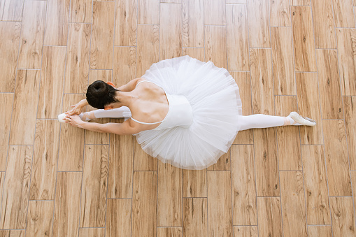 Beautiful graceful Caucasian ballerina in pointe shoes at white wooden floor background, top view from above with copy space. Ballet practice and stretching. Back of ballet dancer.