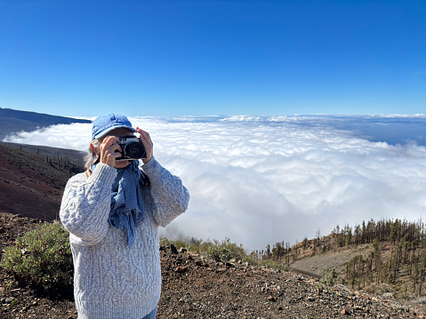 Woman tourist visiting the island of Tenerife takes photographs from the top of the mountain above a sea of clouds, senior female enjoying freedom and beauty in nature