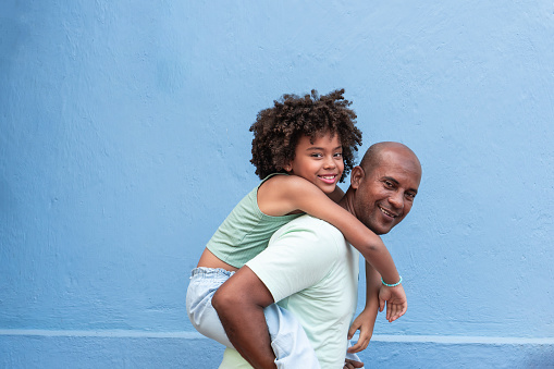 A joyful father carries his young daughter on his back, both smiling against a serene blue background.