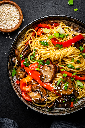 Vegan stir fry noodles with red paprika, champignons, green onion and sesame seeds with ginger, garlic and soy sauce.. Black table background, top view