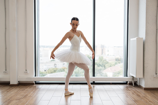 Young ballet dancer - Harmonious pretty woman with tutu posing in studio - Contemporary dance performer. Beautiful ballerina practice ballet positions in white skirt near large window in white hall.