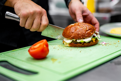 A chef deftly slices a freshly made burger with precision on a green cutting board. Tomato slices await nearby