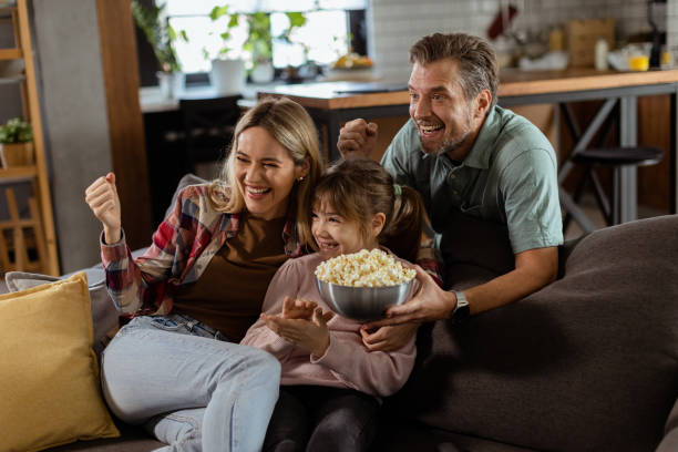 Family Movie Night: Engrossed in Thrilling Scenes at Home A family of three is comfortably nestled on a couch, their faces reflecting excitement and attentiveness as they share a bowl of popcorn during a suspenseful movie night suspenseful stock pictures, royalty-free photos & images