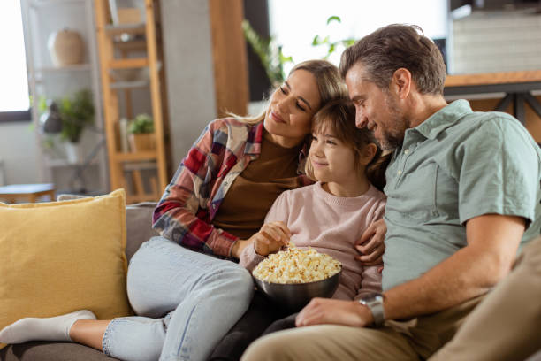 Family Movie Night: Engrossed in Thrilling Scenes at Home A family of three is comfortably nestled on a couch, their faces reflecting excitement and attentiveness as they share a bowl of popcorn during a suspenseful movie night suspenseful stock pictures, royalty-free photos & images