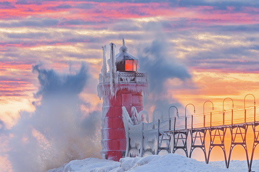 Winter landscape at sunset of the iced South Haven, Michigan lighthouse, catwalk, and pier with splashing wave, Lake Michigan, USA