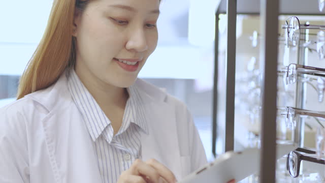 Attractive young female doctor using digital tablet to checks stock of eyeglasses on a shelf in ophthalmology ,eyeglasses store, optical shop. SME, Startup, Owner business.