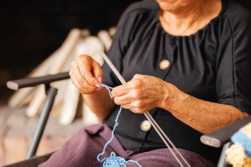 A delightful elderly woman enjoys her retirement, knitting at home with needles and yarn. Her Asian features stand out as she creates diverse knits and does some mending, immersing herself in the creative serenity of her home.