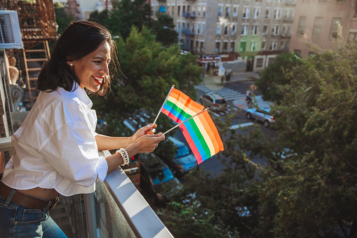 Woman attending to a lgbtqia+ street parade waving flags from her house in New York.