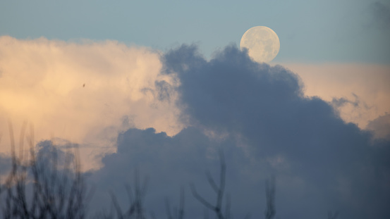 Full moon shining on an early morning, with part of it covered by white and dark clouds.
