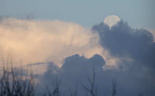 Full moon shining on an early morning, half is swallowed by dark clouds, positioned on the right, left side are white clouds