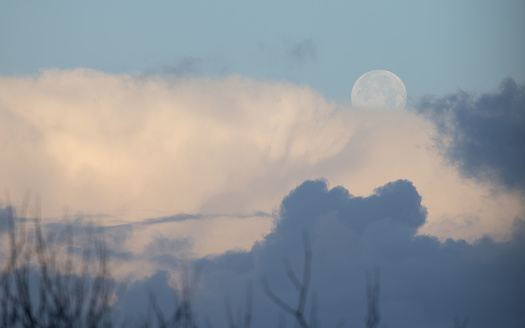 Full moon shining bright on an early morning, illuminated, with white clouds underneath.