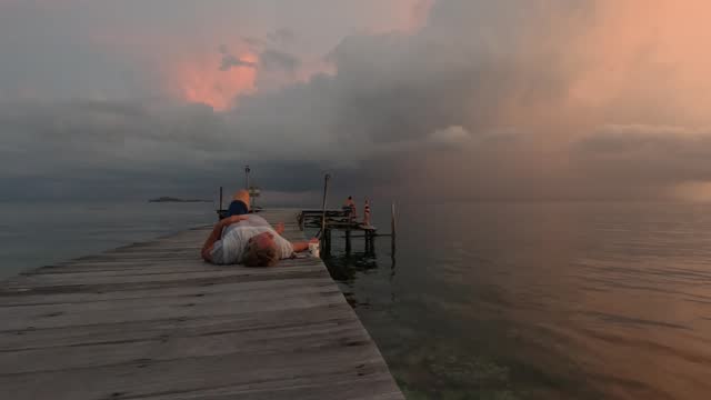 An adult man lies on a boardwalk pier and admires a magnificent bright sea sunset, West Papua, Indonesia