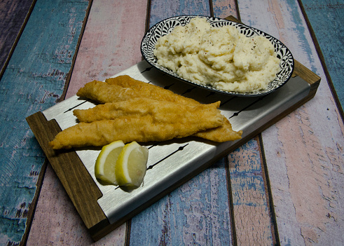 Closeup view of fried breaded fish filets with lemon and potato puree in a metal board on the wooden table.