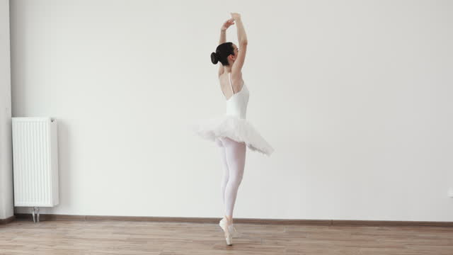 Caucasian woman ballerina in white tutu, dancing on pointe with arms overhead.