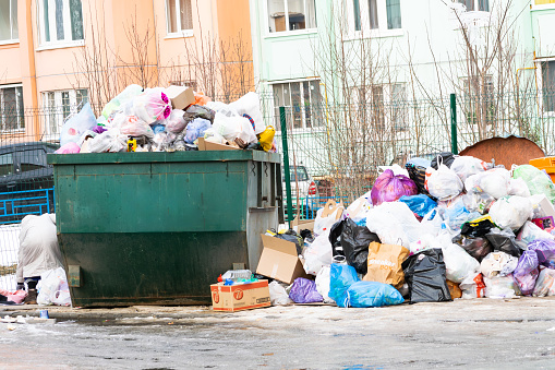 Moscow, Russia - March 03, 2024: a garbage container site littered with unsorted household waste and a poor anonymous person rummaging through it in search of food. Garbage not removed on time